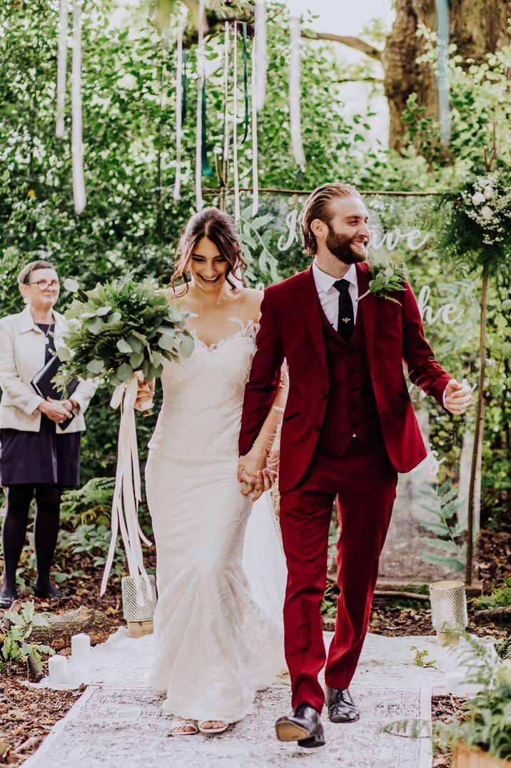 Outdoor woodland wedding with bride in a lace wedding dress and groom in a burgundy red wedding suit descending up the aisle as husband and wife