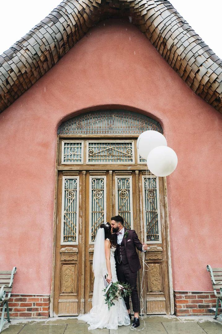 Groom in tweed suit and bride in lace wedding dress holding balloons standing outside Le Petit Chateau 