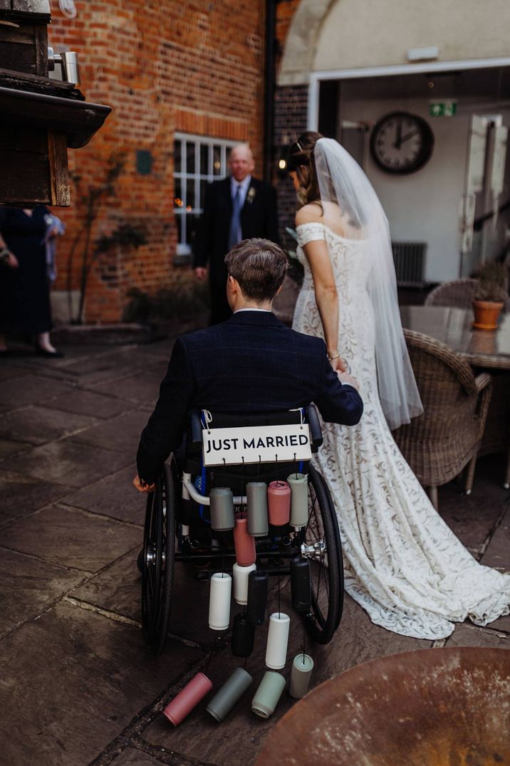 Bride in lace off shoulder dress with veil and groom in dark suit in wheelchair with DIY Just Married sign and colourful painted tin cans decorations