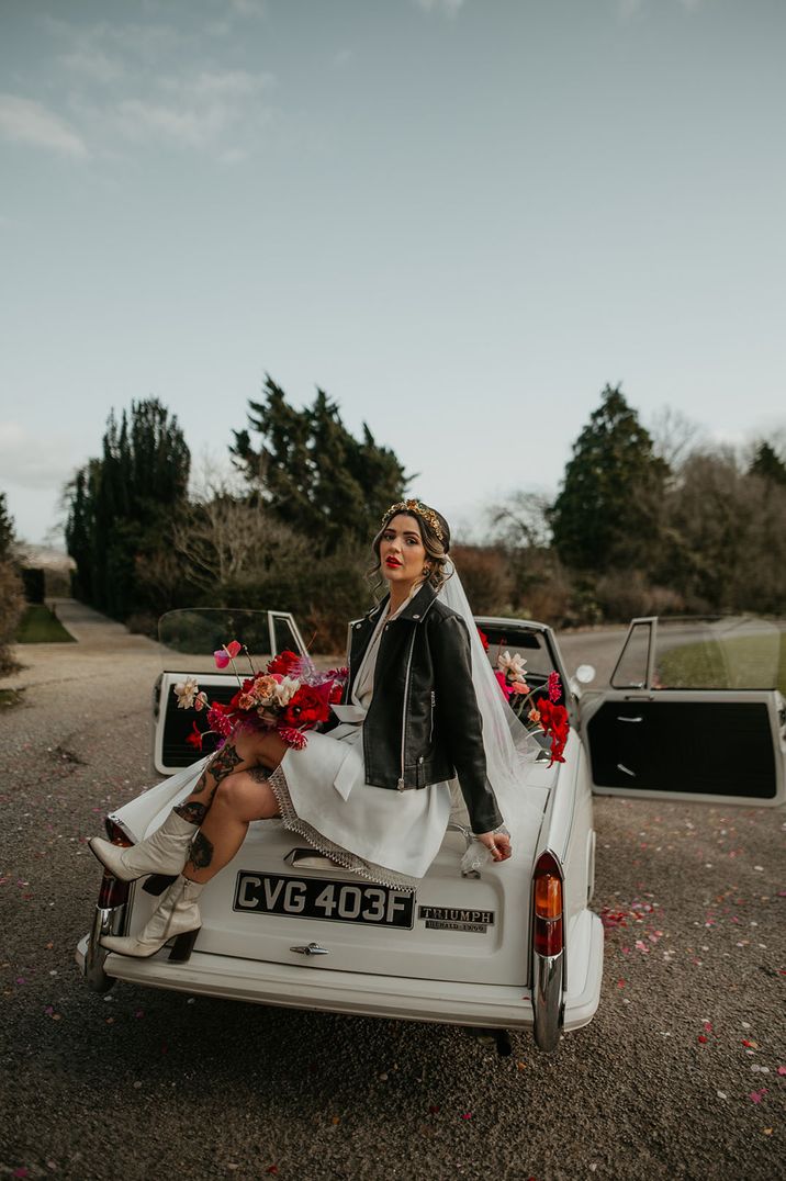 Bride with white boots sitting on white wedding car with black leather jacket holding vibrant bouquet 