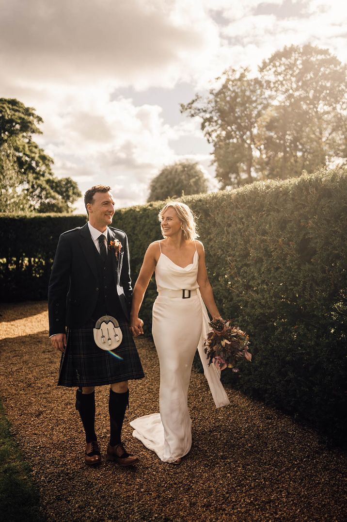 Bride walking along hand in hand with the groom in a satin slip wedding dress with cowl neckline and belt accessory 