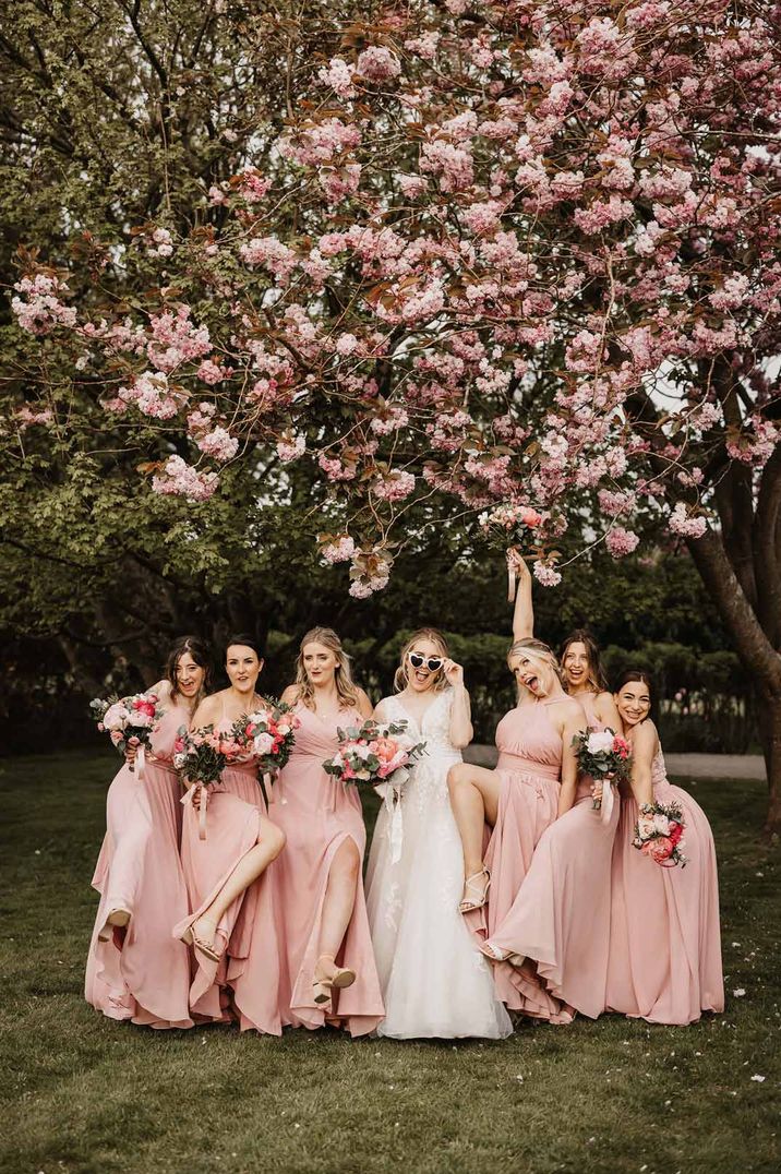 Bride in off shoulder lace wedding dress posing with bridesmaids in mismatched baby pink bridesmaid dresses underneath pink cherry blossom tree