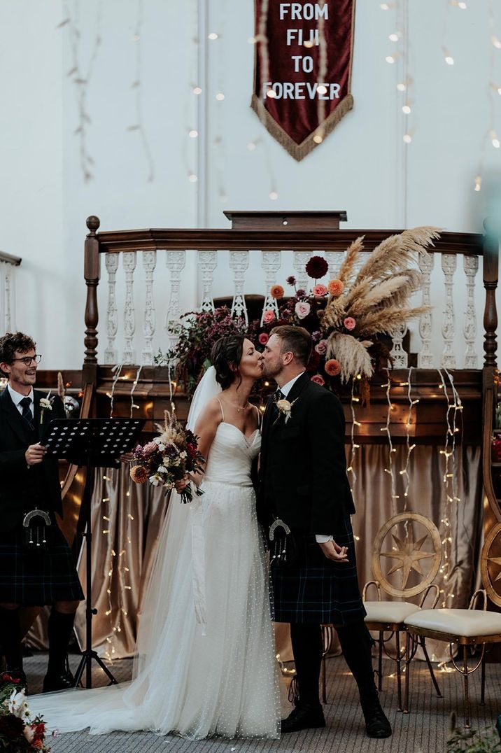 bride in a polka dot tulle wedding dress overlay kissing her groom in a brown suit at the altar decorated with dried flowers and wool rugs