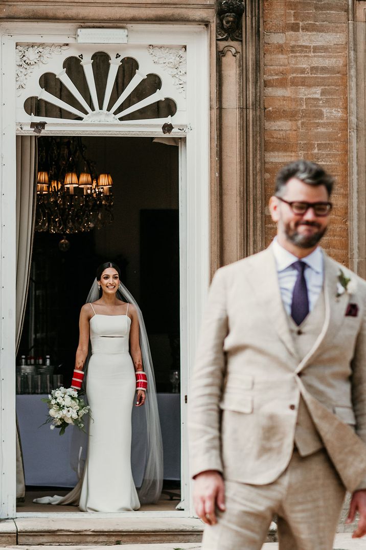 Bride in fitted wedding dress approaches groom from behind for private first look photographs 