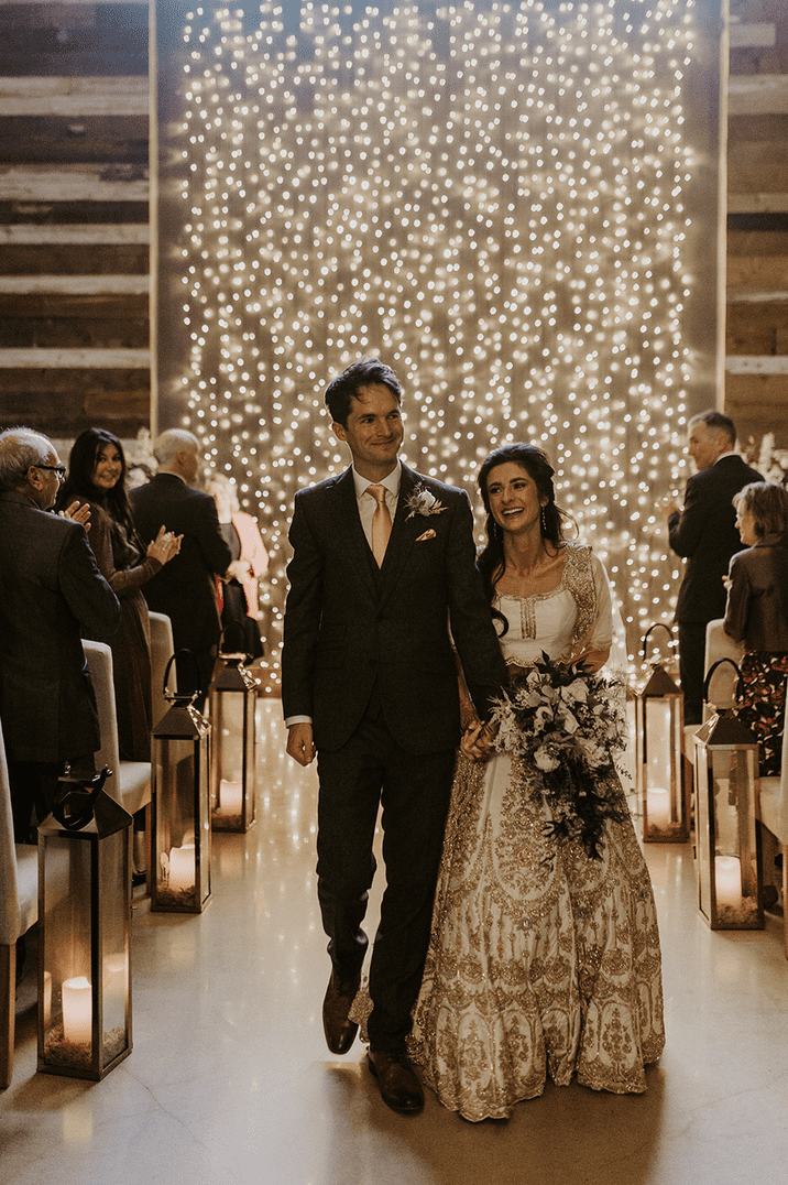 The bride and groom walk back down the aisle together with a fairy light backdrop 