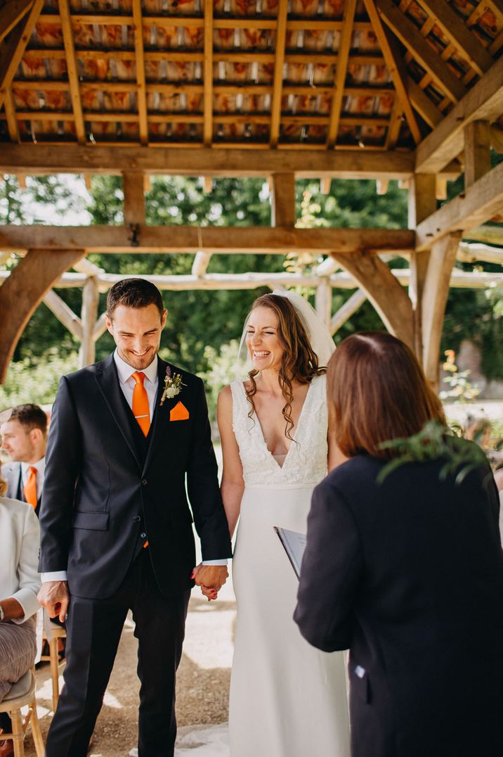 bride in plunging neck line fitted wedding dress laughing with her husband in a navy suit and orange tie at their outdoor wedding ceremony 