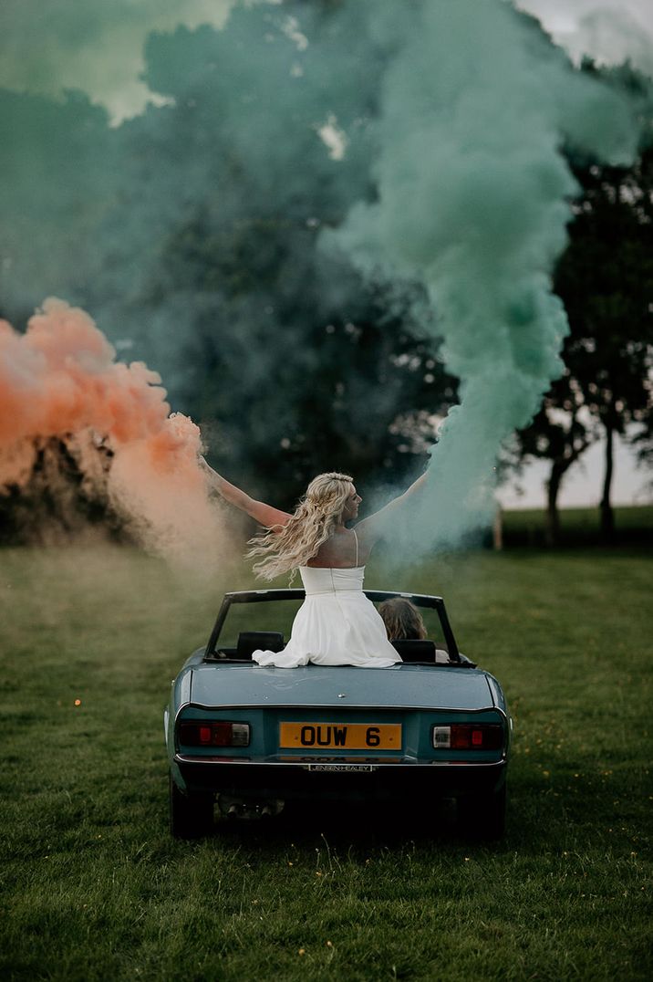 Bride sitting on the back of a convertible car holding an orange and green smoke bomb in each hand 