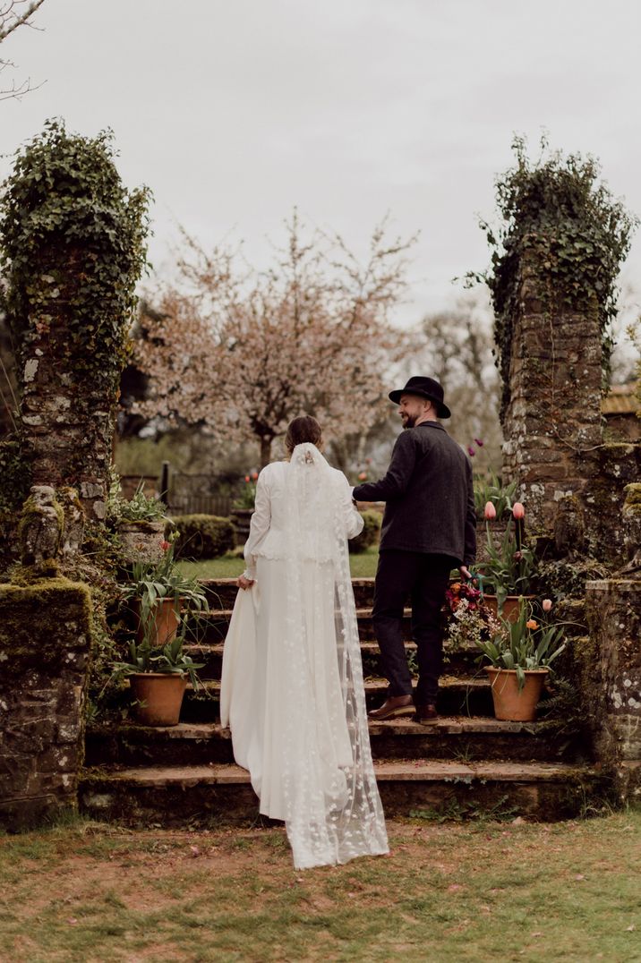 Garden wedding with groom in a fedora hat holding hands with his bride in an appliqué veil 