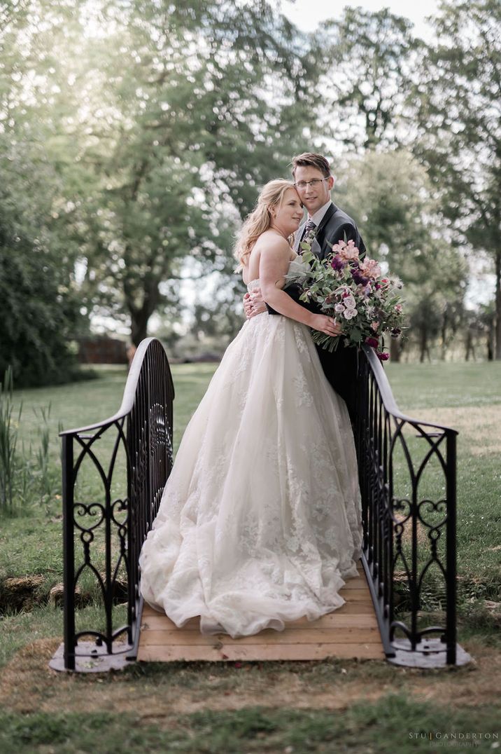 Bride in princess wedding dress with pink and purple wedding bouquet embracing the groom on a bridge for 60k wedding 