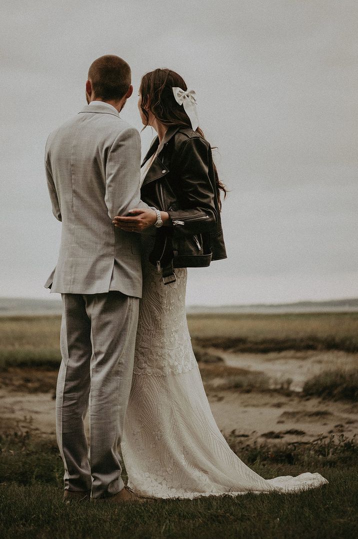 Bride in beaded wedding dress with black leather jacket wearing hair bow accessory with the groom in light suit 