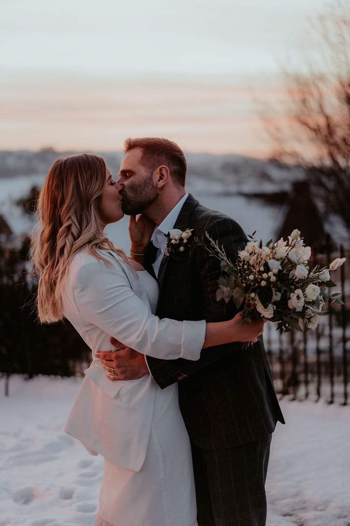 Bride wearing white blazer jacket kisses the groom during sunset at snowy winter wedding 