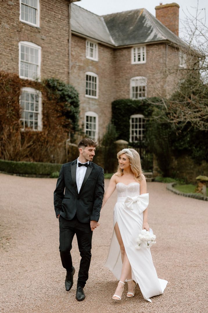 Bride in sparkly corset wedding dress with bow detail walks with the groom in a black tux at West Midlands wedding venue 