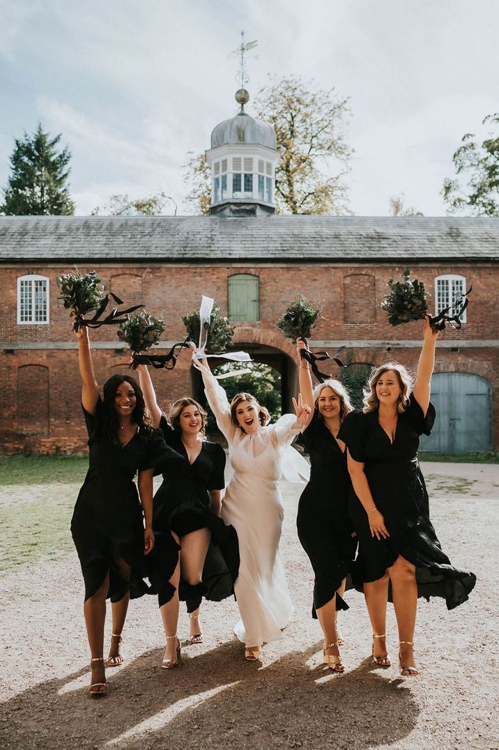 Bridesmaids in black dresses raise their preserved all foliage bouquets walking with the bride 