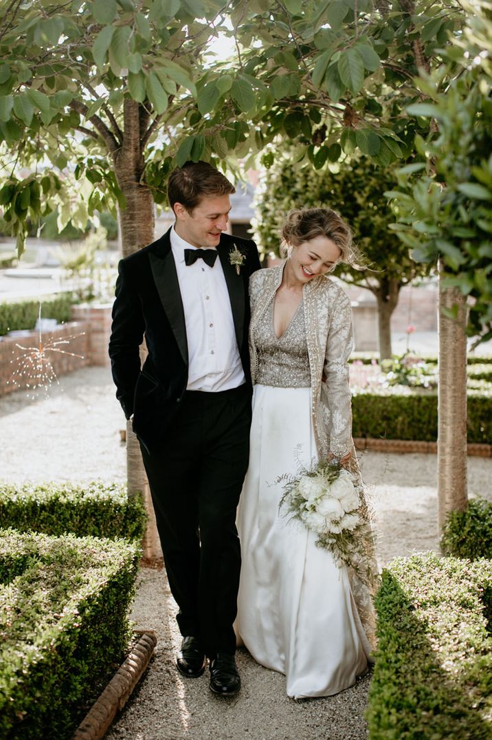 Groom in black tuxedo walking along with bride in gold embellished top, white skirt, and Indian bridal coat 