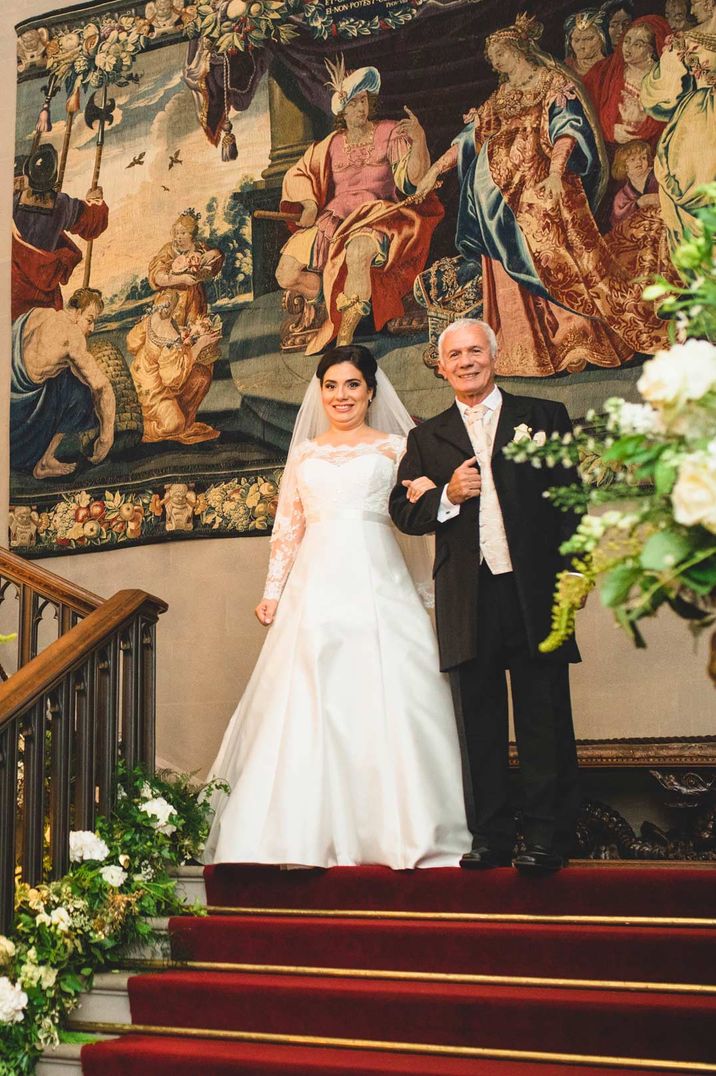 Bride in long sleeve lace wedding dress with scoop neck and short veil walking down the red carpeted stairs of Eastnor Castle with large religious painting and foliage, dried flower, peony and garden rose flower arrangements 