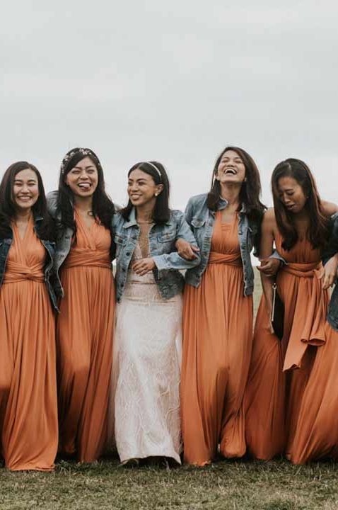 Bridesmaids in front wrap orange bridesmaid dresses and dark wash denim jackets walking with bride in lace wedding dress with denim bridal coverup