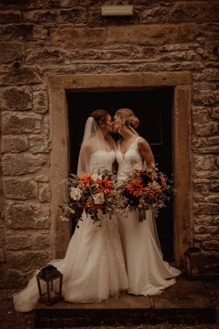 Two brides share a kiss in embellished lace wedding dresses holding matching floral bouquets 