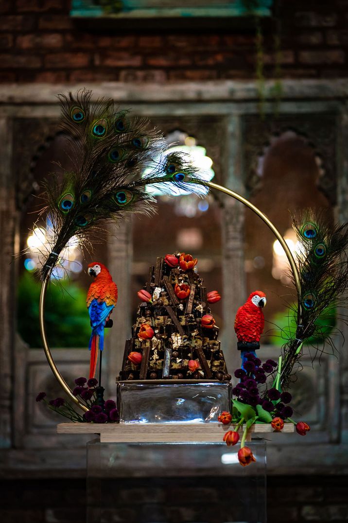 Chocolate wedding cake with chocolate biscuit fingers and red tulip decor on a hoop cake stand decorated with peacock feathers and parrots 