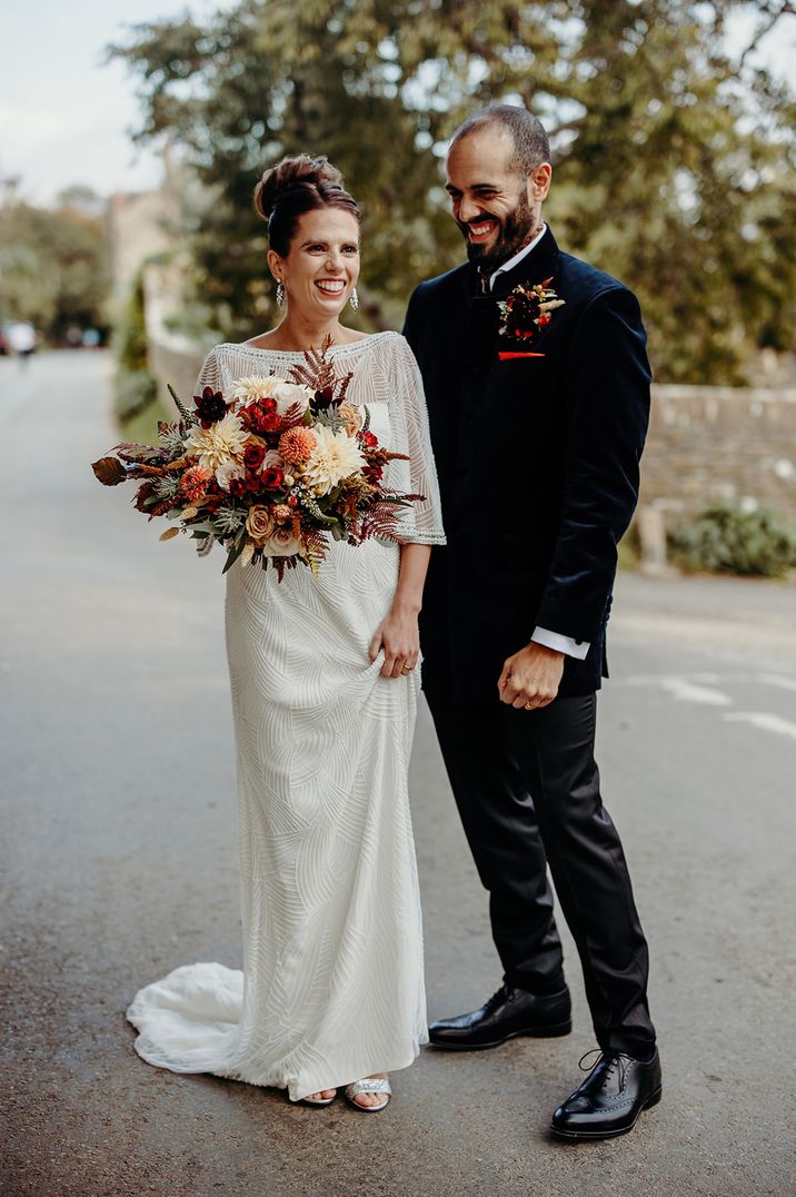 Bride in an embellished wedding dress holding an autumn wedding bouquet with dahlias laughing with her husband in a navy velvet jacket 