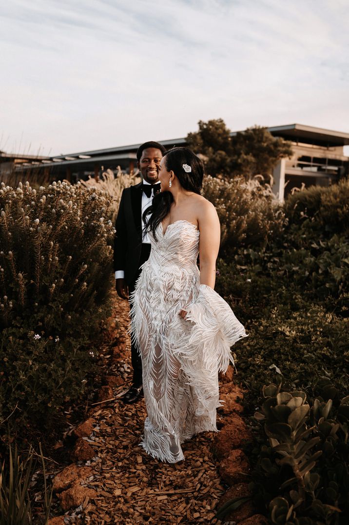 Bride in a couture strapless wedding dress leading her Black groom in a tuxedo through the fields