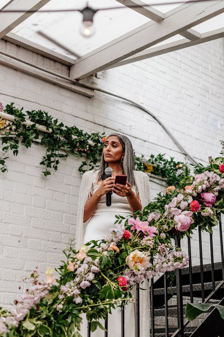 Bride makes wedding speech in long white wedding dress with fringed shall on floral staircase