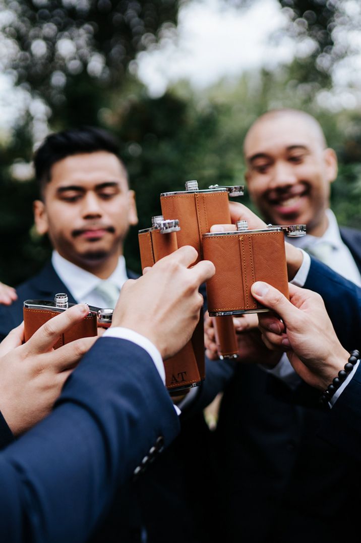 Grooms in dark blue suits toasting with brown leather hip flasks