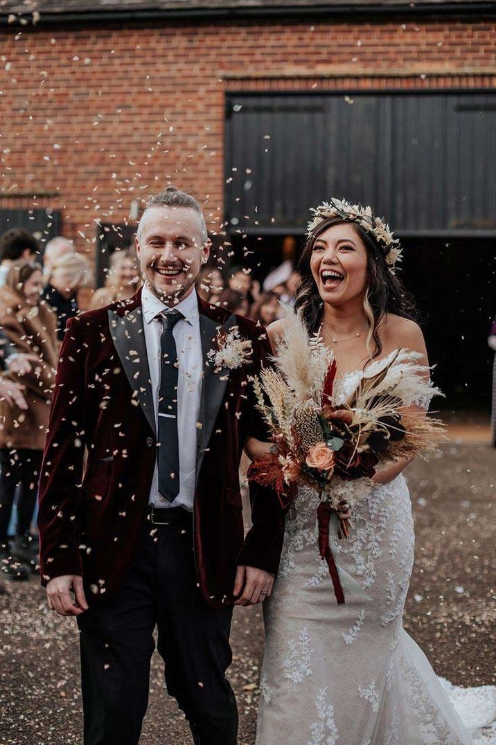 Bride in lace wedding dress and bridal flower crown doing confetti exit with groom in classic black tuxedo