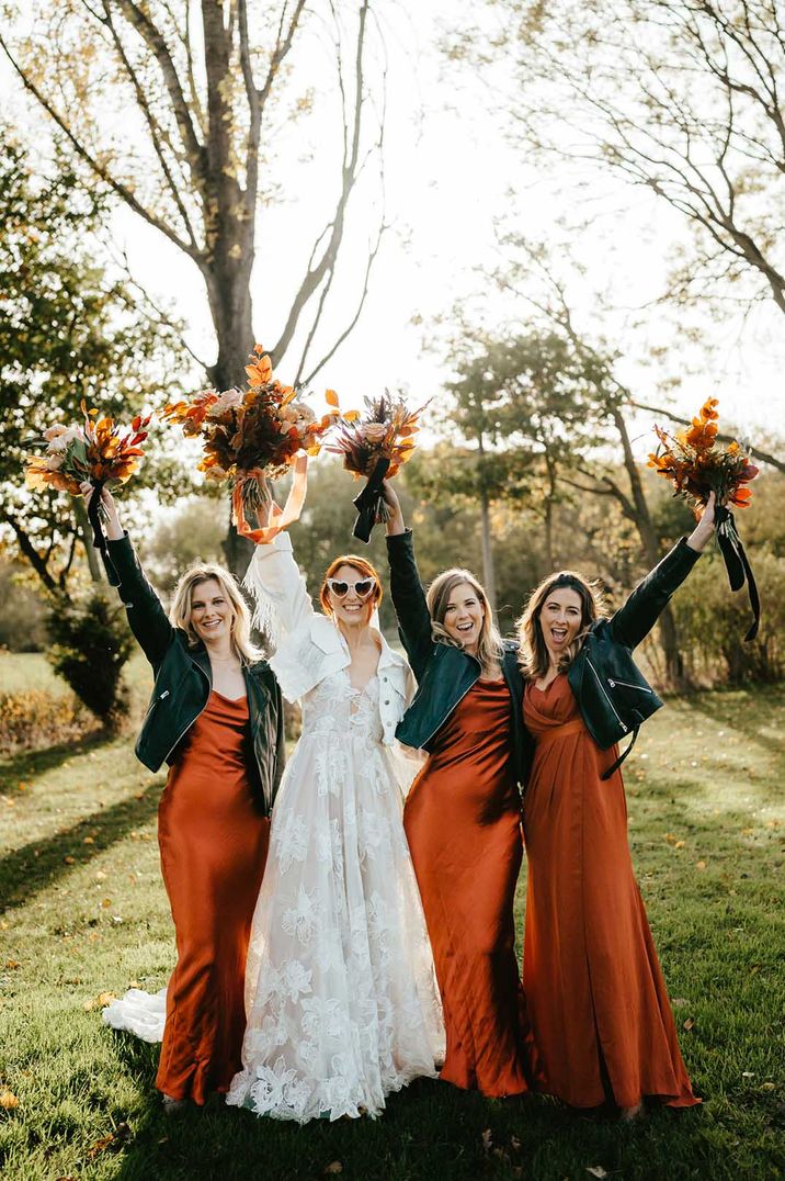 Bride in 3d applique leaf wedding dress with white denim jacket bridal coverup standing with bridal party in burnt orange bridesmaid dresses and black cropped jackets 