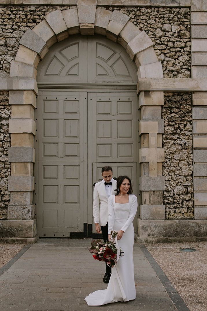 Groom in white tuxedo waits behind the bride in a sparkly wedding dress for first look wedding photos 