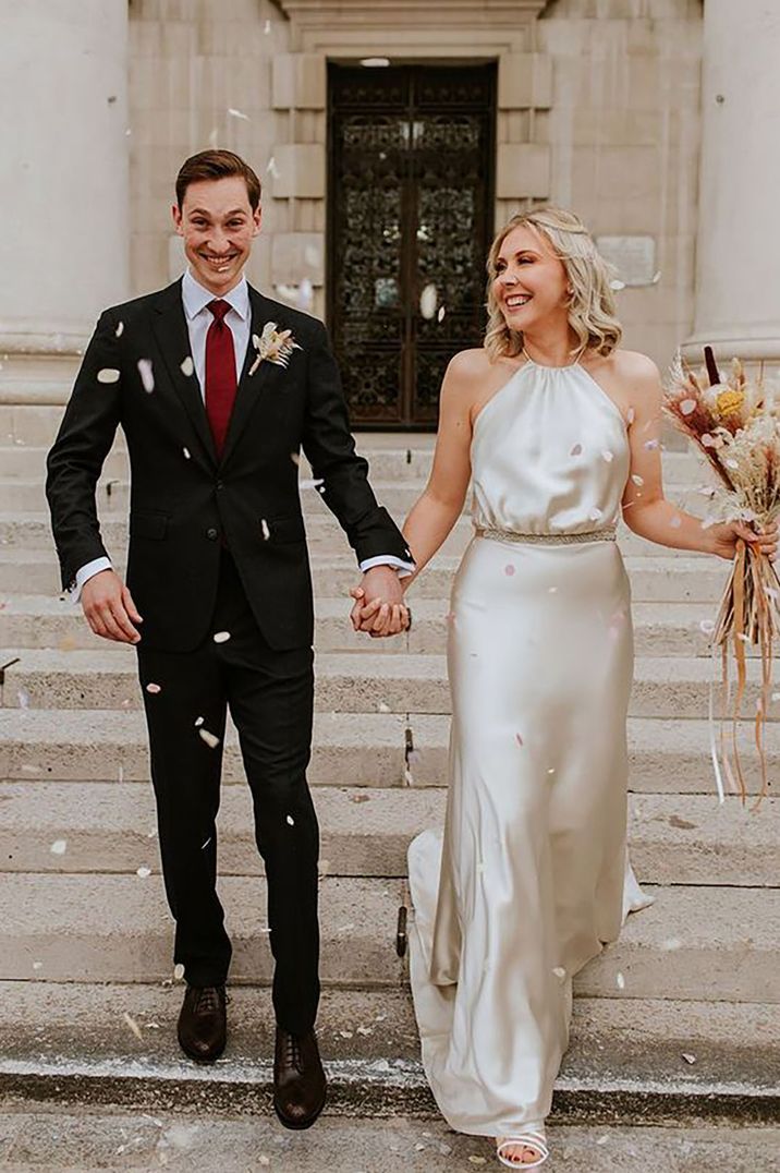 Bride wearing a satin halterneck dress and groom in black suit coming down the stairs with confetti at registry office wedding