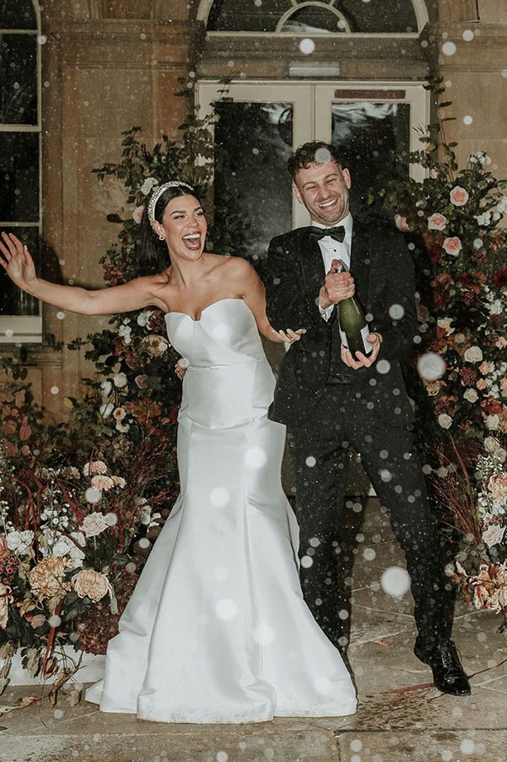 Bride in mermaid wedding dress with groom in black tuxedo popping bottle of champagne 