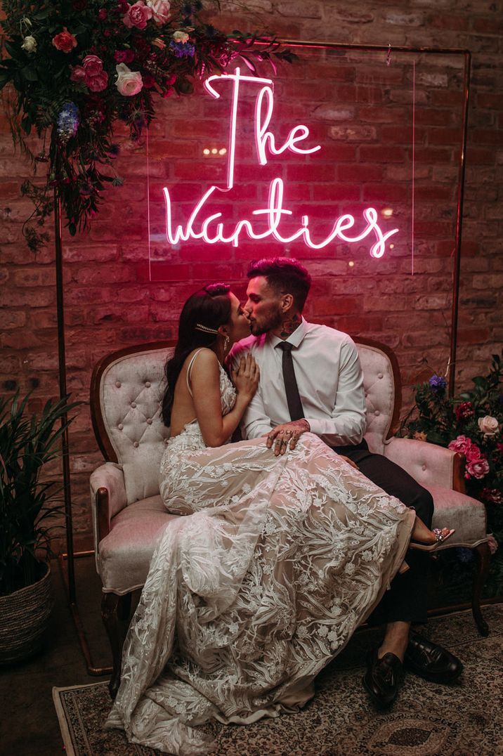 Bride in a lace wedding dress cosying up to her husband on an ornate sofa under a personalised pink neon wedding sign 