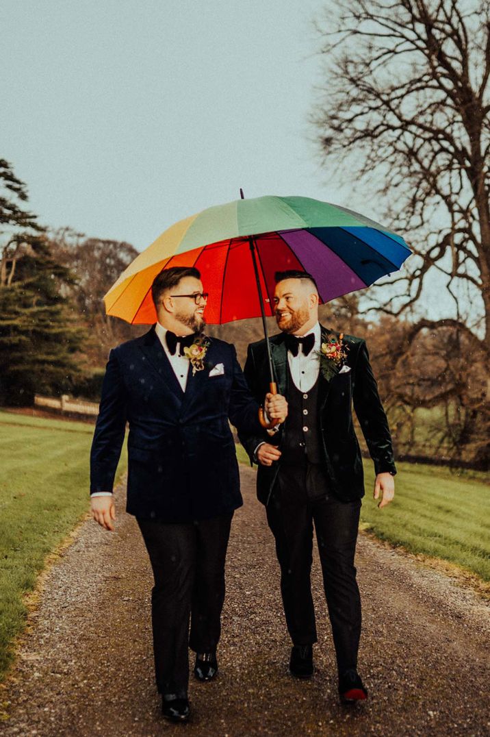 Grooms walk under rainbow coloured umbrella in velvet suit jackets