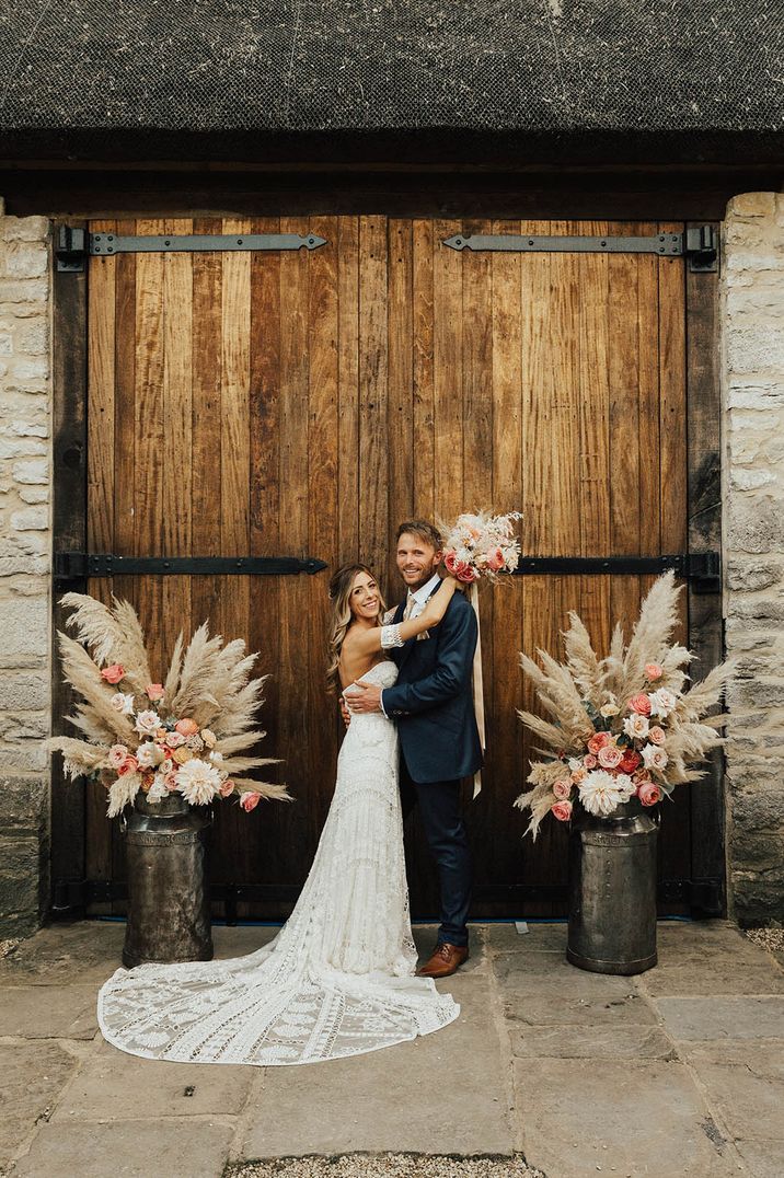 Epic pink and white rose and pampas grass in milk churns with the bride in a boho strapless lace wedding dress and groom in navy suit 