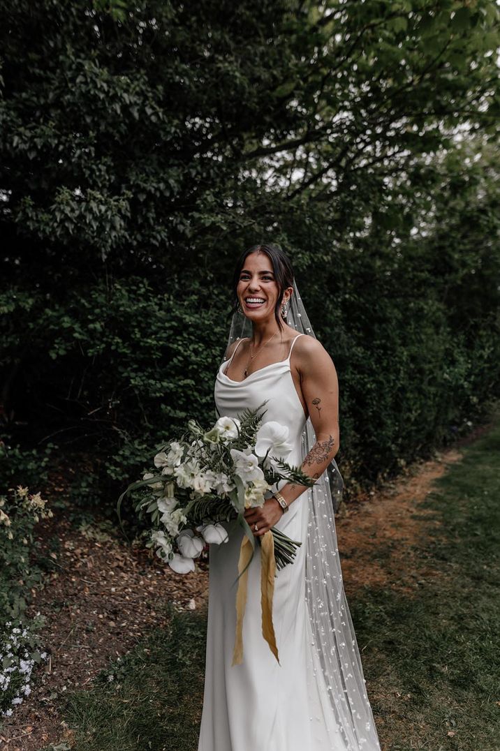 Bride in satin cowl neck wedding dress and pearl veil with white flower wedding bouquet for chic outdoor wedding 