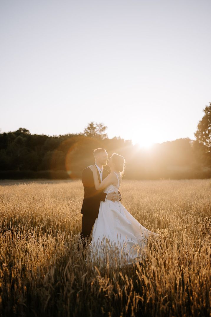 Groom in black suit jacket embraces the bride in a wedding dress standing in a wide open field with pretty sunset 