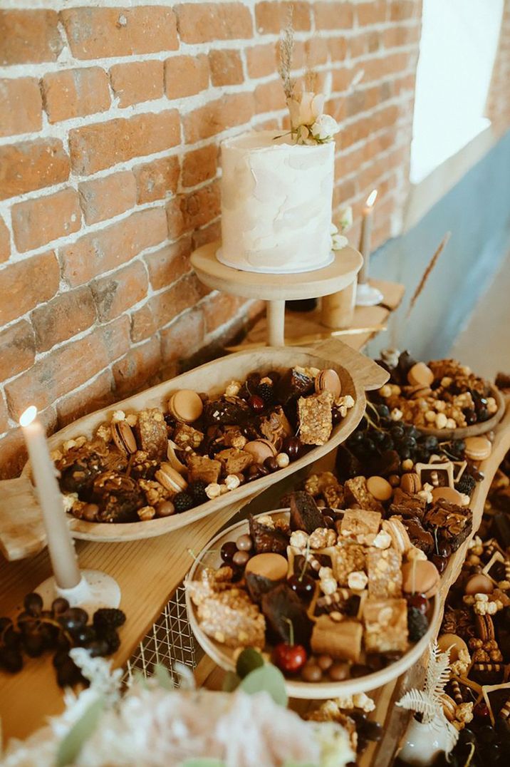 Wedding dessert table with trays of flapjacks, brownies, popcorn, macarons and more with a single tier white iced wedding cake 