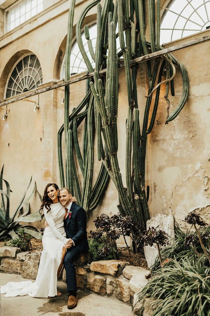 Bride in long sleeve wedding dress and groom in dark suit sitting and embracing next to large cacti installation at Syon Park, London glass house wedding venue 