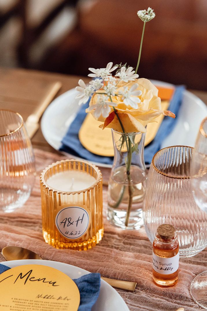 Personalised wedding stationery at rustic wedding tablescape with blue cotton napkins, yellow candle holder, and yellow rose 