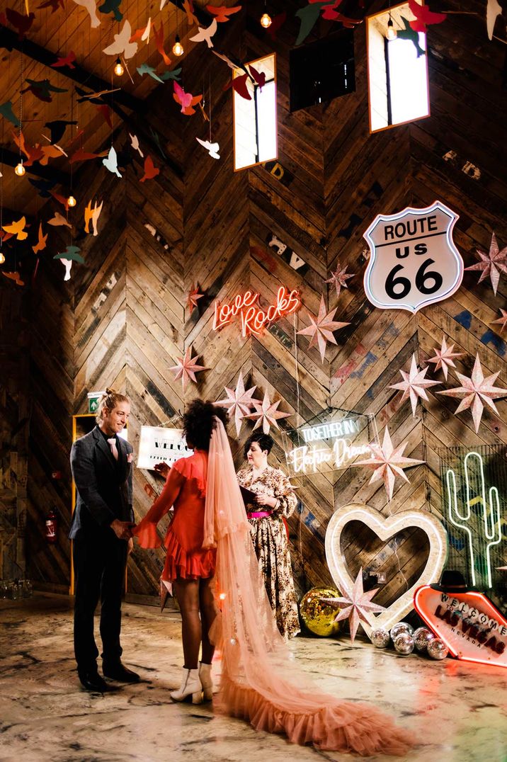 Bride in long sleeve short red wedding dress and pink two tier veil holding hands with groom in dark suit with an abundance of neon wedding signs 