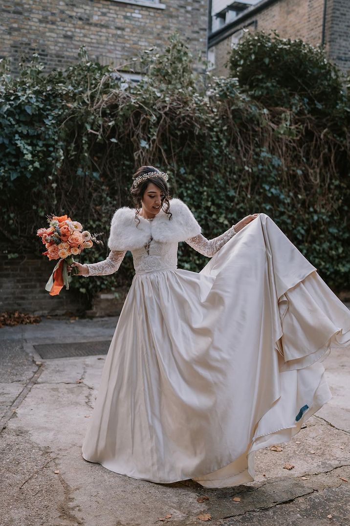 Bride in princess wedding dress with a faux fur shawl and crown holding a pretty bouquet of pink, peach, and orange flowers 