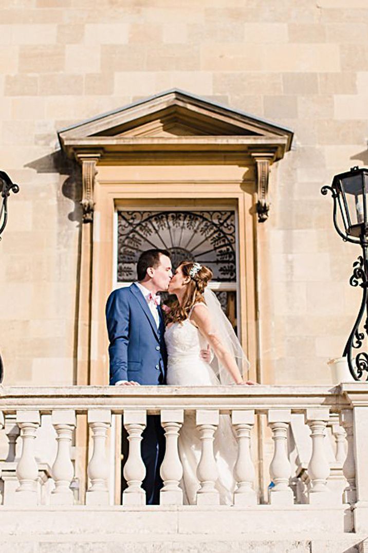 Bride in strapless peplum wedding dress kissing groom in blue suit and pink bowtie standing on the balcony of Botleys Mansion - one of the wedding venues London 