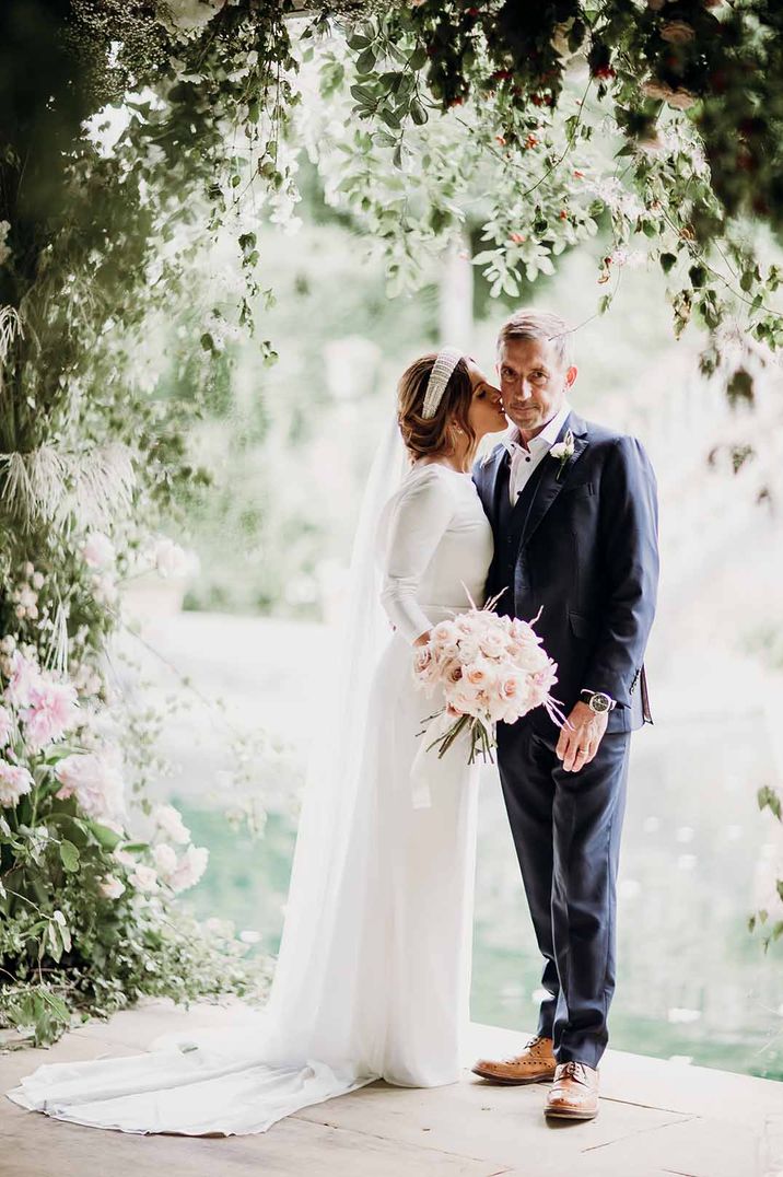 Elbeth Gillis wedding dress worn by bride with pearl headband and pink rose bouquet standing with the groom in navy suit