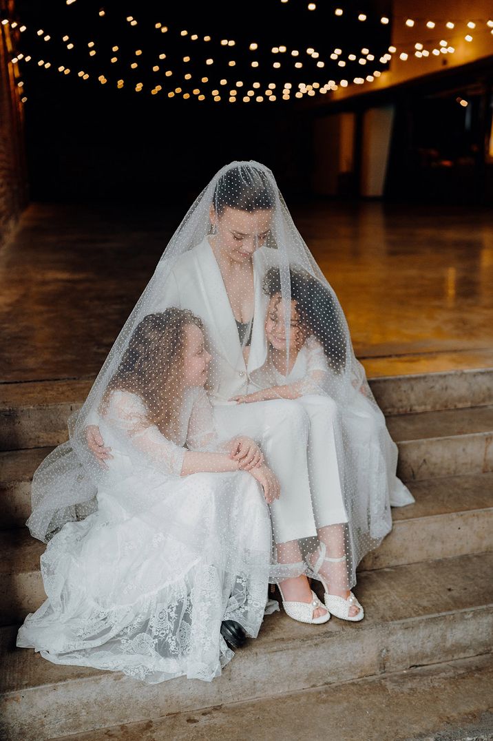 The bride sits in her white bridal suit with dotted veil covering her and her two daughters 