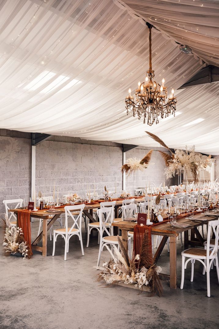 Boho wedding reception decor with draped ceiling and fairy lights, pampas grass flowers and rust coloured table runners 