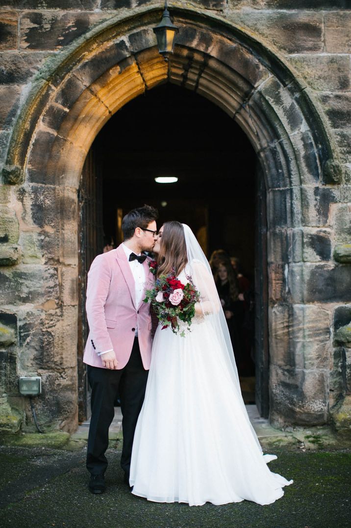 Groom in a pink velvet suit jacket kisses the bride for their winter church wedding 