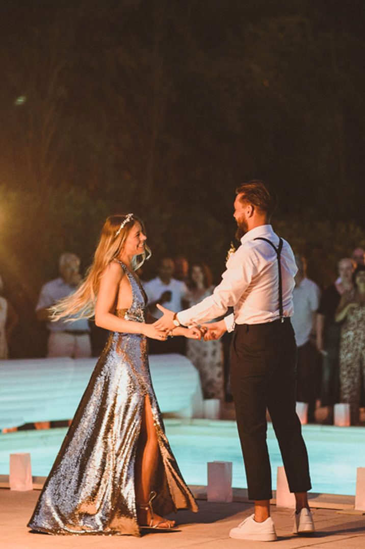 The bride wears a long silver metallic wedding reception dress dancing with the groom next to a pool at their destination wedding 