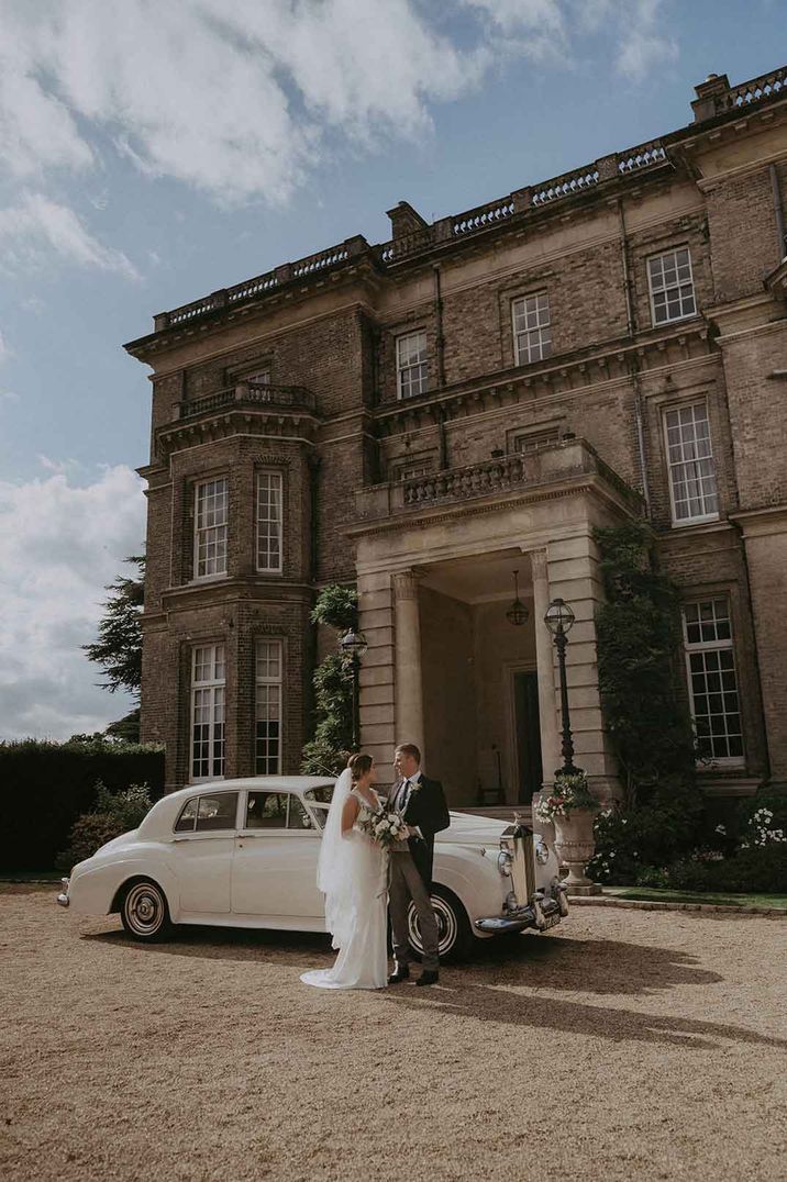 Bride in Savannah Miller wedding dress and groom in three piece grey suit standing by classic wedding car outside of Hedsor House country house wedding venue search