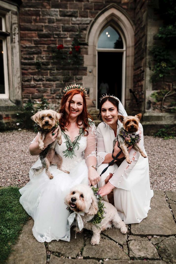 Brides in long sleeved sheer v neck wedding dresses with gold bridal crowns holding three puppies on the grounds of Dunwood Hall Estate castle wedding venue