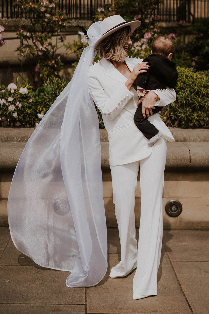 Bride in pearl Nadine Merabi suit with a cream hat and veil attached holding a baby 